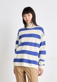 Sweter w kremowym kolorze z wyraźnymi niebieskimi poziomymi paskami, projekt z kołnierzem w kształcie lejka, luźny krój, ściągacze z prążkowanej dzianiny i subtelny detal z logo na dole.