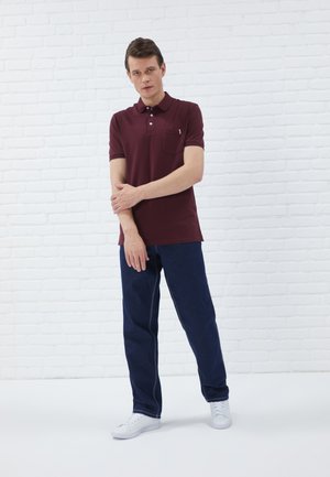 Bordeaux polo met een kraag en drie knopen, gecombineerd met donkerblauwe jeans. Het model draagt witte sneakers. Gestructureerde achtergronden.