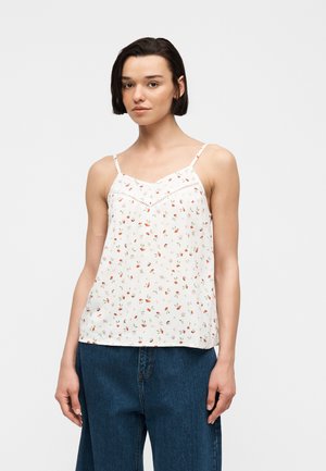 Kvinde med kort mørkt hår iført en hvid blomstret top med spaghetti-stropper og blå cowboybukser, stående mod en ensfarvet baggrund.