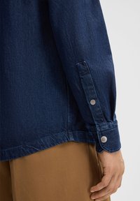 Denimjack, donkerblauw, met metalen drukknoopjes op de mouwen. Heeft een rechte zoom en een stevige stoftextuur. Close-up van de mouw en de manchet.