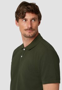 Donkergroene polo met een platte kraag, korte mouwen en drie knopen aan de voorkant. De stof lijkt glad met een klassieke pasvorm.