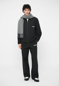 Schwarze Zip-Jacke und passende Hose mit "Double"-Logo. Grauer Fransen-Schal. Glatte Textur, moderne Passform, schlichtes Design, keine sichtbaren Muster.