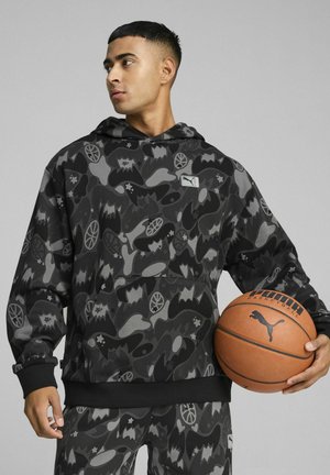 Mustmust ja halli mustriga hoodie, mis on valmistatud pehmest kangast, millel on kengurutasku ja PUMA logo päts. Modell hoiab korvpalli.