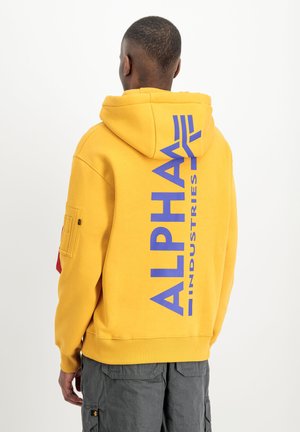 Mann trägt gelben Hoodie mit großem vertikalem blauem Schriftzug "ALPHA INDUSTRIES" auf dem Rücken, kombiniert mit grauen Cargohosen, steht mit dem Rücken zur Kamera.