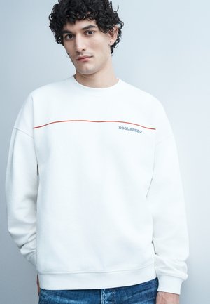 Noore mehe lokkis juustega, kes kannab valget DSQUARED2 pusa õhukese punase horisontaalse triibuga, siniste teksadega, seisab halli tausta ees.