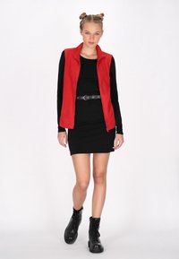 Vesta roșie, fără mâneci, peste o rochie ajustată neagră, accesorizată cu o curea neagră și botine negre cu șireturi până la genunchi. Țesătură netedă și linii curate.
