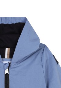 Veste bleu clair avec un col haut, doublure noire, fermeture éclair frontale et un petit patch logo noir sur la manche gauche.