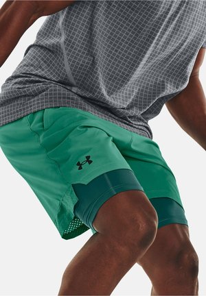 Mann, der grüne, geschichtete Sportshorts und ein graues gemustertes Hemd trägt, in einer hockenden oder aktiven Pose aufgenommen.