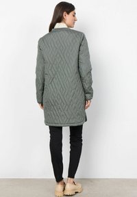 Vadderad lång kappa i olivgrön med ett geometriskt mönster, rundad krage och sidofickor. Bärs över svarta byxor och beige skor.
