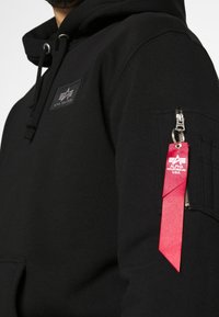 Felpa nera con cappuccio, tasca frontale, coulisse e patch con logo. Presenta una tasca con zip sulla manica con un'etichetta rossa e hardware argentato.