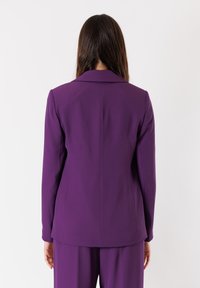 Blazer viola con spalle strutturate, spacchetto posteriore singolo e tessuto liscio. Design minimalista, maniche lunghe e colletto a revers classico.