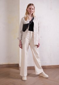 Giovane donna in piedi dentro casa con giacca a righe, top nero, pantaloni larghi crema, sneakers bianche e borsa a tracolla nera.