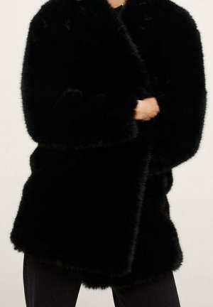 Personne portant un manteau noir en fausse fourrure duveteuse à manches longues, associé à un pantalon noir, debout devant un fond clair uni.