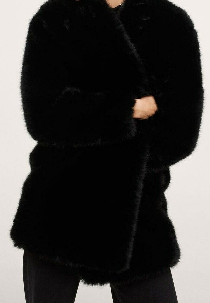 Personne portant un manteau noir en fausse fourrure duveteuse à manches longues, associé à un pantalon noir, debout devant un fond clair uni.