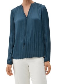 Blusa plissettata color teal con scollatura a V, maniche lunghe e dettagli argento nei nastri, realizzata in un tessuto liscio e lucido. Abbinata a pantaloni bianchi.