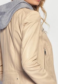 Tan läderjacka med grå huva. Har sidofickor med dragkedja och detaljerad sömnad. Slät textur med en åtsittande design.