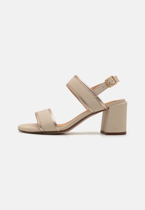 Beige heeled sandals with metallic gold accents, featuring wide straps and a block heel design. Smooth texture and ankle buckle closure.