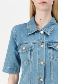 Jaqueta de ganga azul claro com fecho de botões à frente, dois bolsos no peito com botões em tom de ouro e colarinho pontudo. Mangas curtas.