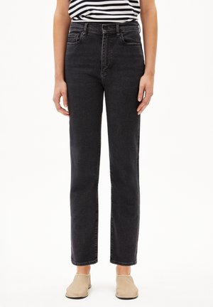 Personne portant un jean noir taille haute à jambes droites, un top sans manches rayé noir et blanc, et des chaussures beige slip-on, debout sur fond blanc.