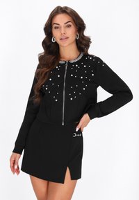 Chaqueta negra recortada con detalles de perlas y cremallera plateada, combinada con una minifalda negra que presenta una abertura lateral y detalles metálicos.