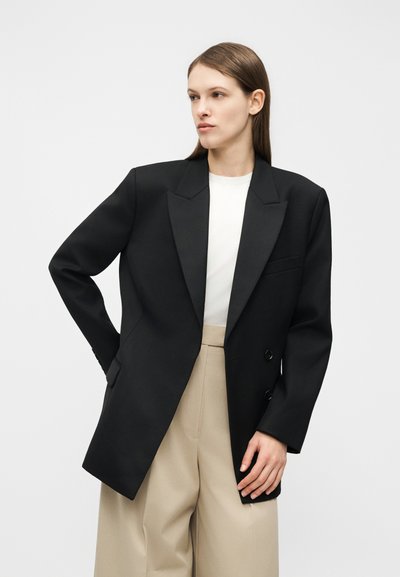 Žena v prehnane veľkom čiernom dvojradovom blejzri cez biely top a voľné široké béžové nohavice, stojaca s jednou rukou na páse.