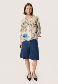 Blusa floreale trasparente con motivi blu, verdi e marroni, abbinata a pantaloni corti in denim blu e décolleté nere a punta.