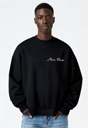 Jeune homme debout, les mains dans les poches, portant un sweat-shirt noir avec le texte « Mira Paris » et un jean bleu clair, fond neutre.
