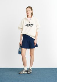 Krémová mikina s čiernym logom "ADIDAS", námornícke plisované sukne, šedé topánky s čiernymi pruhmi a malou modrou kabelkou.