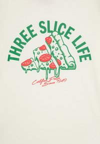 Krém színű póló, amelyen három, piros feltéttel rendelkező pizzaszelet grafika látható, zöld felirattal: "HÁROM SZELET ÉLET," és piros díszítésekkel.