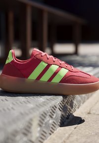 Red sneaker with neon green stripes and gum sole resting on textured metal surface in outdoor setting.