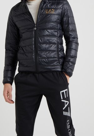 Hombre con chaqueta acolchada negra brillante con el logo EA7 Emporio Armani y pantalones negros con el gran logo blanco EA7 en la pierna izquierda.