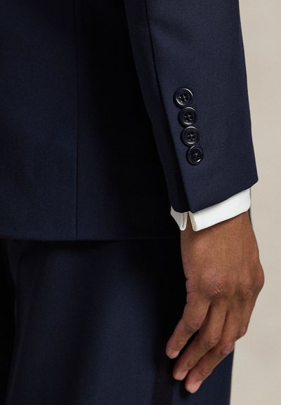 Tamsiai mėlyno kostiumo rankovė su lygiu paviršiumi, keturiais juodais mygtukais ir baltu rankogaliu, išlendančiu iš po švarko.