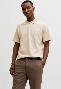 Homme portant un polo beige à manches courtes avec une fermeture éclair à l'avant et un pantalon marron, debout avec une main dans la poche devant un fond uni.