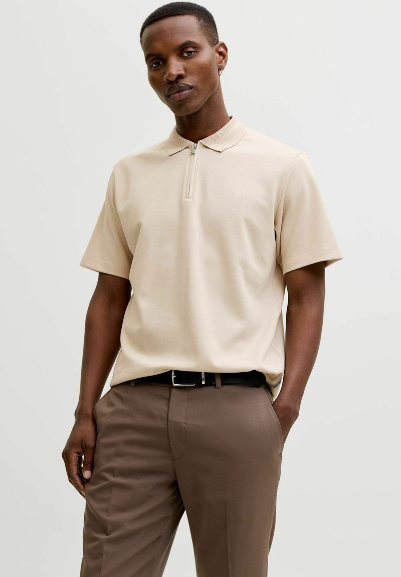 Homme portant un polo beige à manches courtes avec une fermeture éclair à l'avant et un pantalon marron, debout avec une main dans la poche devant un fond uni.