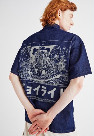Camicia di jeans a maniche corte di colore blu scuro con una stampa grafica sul retro che presenta disegni intricati e elementi testuali bianchi.