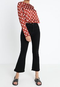 Rotes Satin-Oberteil mit langen Ärmeln und weißen Punkten, kombiniert mit schwarzen Schlagjeans und schwarzen Sandalen mit weißen Punkten.
