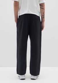 Schwarze weit geschnittene Hosen aus weichem Stoff, mit einer Gesäßtasche und subtiler Markenkennzeichnung an der Seite. Kombiniert mit einem schlichten weißen Hemd und Sneakers.