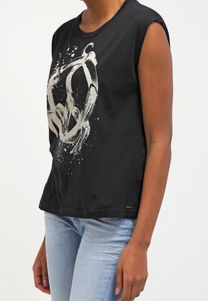 T-shirt noir sans manches avec un graphisme abstrait blanc, porté par une personne en jean bleu clair, debout devant un fond clair uni.