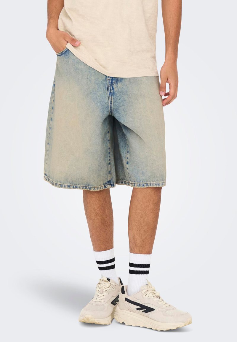Svetlomodré denim šortky s voľným strihom, surovým lemom a viditeľnými detailami stehov. Nosí sa s bielymi ponožkami so čiernymi pruhmi a teniskami.