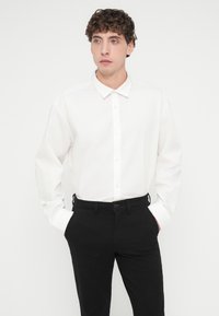 Chemise blanche à manches longues avec col classique et boutons, associée à un pantalon noir à devant plat et passants pour ceinture. Tissu à texture lisse.