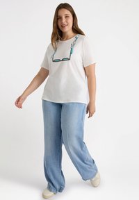 Hvid kortærmet t-shirt med et mønstret detalje ved halsudskæringen, parret med lyseblå vide jeans og beige sko. Glat stof og afslappet pasform.