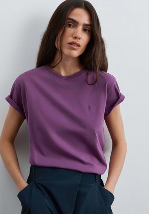 Mujer con cabello largo y oscuro lleva una camiseta morada con las mangas remangadas y pantalones oscuros, de pie frente a un fondo liso gris claro.