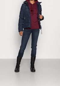 Námořnická modrá péřová bunda, se zavíráním na zip, nošená přes vínový hoodie s nápisem. Tmavě modré džíny a černé šněrovací boty doplňují celý outfit.