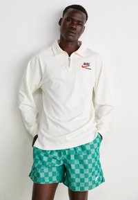 Hvit langermet zippered genser med Nike-logo, kombinert med grønne rutete svømme shorts med en liten logo på kanten.