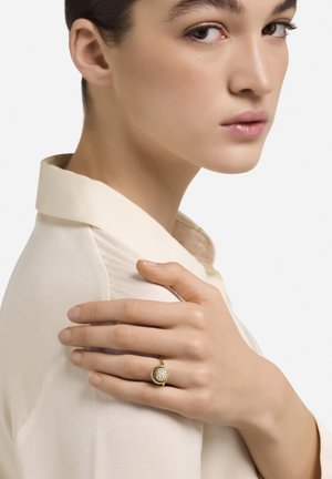 Zlati prstan z okroglim vrhom, okrašenim z majhnimi kristali, ki ga drži roka na bledo svileni bluzi z nizkim ovratnikom.