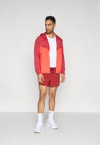 Ελαφρύ μπουφάν σε κόκκινο και πορτοκαλί με κουκούλα, λευκό T-shirt, κόκκινα σορτς, λευκές κάλτσες Nike και αθλητικά παπούτσια. Απεικονίζεται πάνω σε ουδέτερο φόντο.