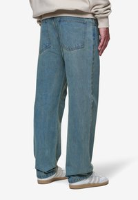 Jeans de ganga azul claro, de perna larga, com cintura alta, apresentando dois bolsos traseiros e desbotamento subtil. Combinados com ténis brancos.