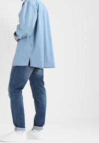 Osoba ubrana w jasnoniebieską koszulę dżinsową, ciemnoniebieskie dżinsy z mankietami, białe skarpetki i białe sneakersy, stojąca na tle białej ściany.