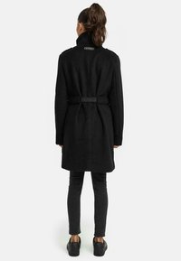 Cappotto di lana nero con colletto alto, vita con cintura e dettaglio del logo sulla schiena. Indossato con jeans skinny scuri e sneaker nere.