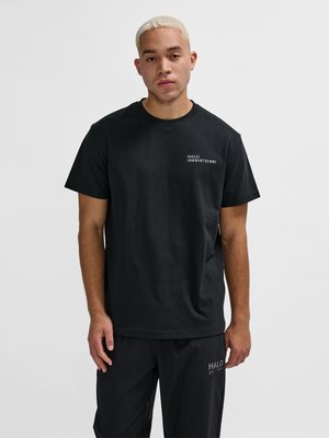 Ung mand med kort blondt hår iført en sort T-shirt og bukser med hvidt "HALO"-tekst på brystet og låret, stående mod en ensfarvet baggrund.